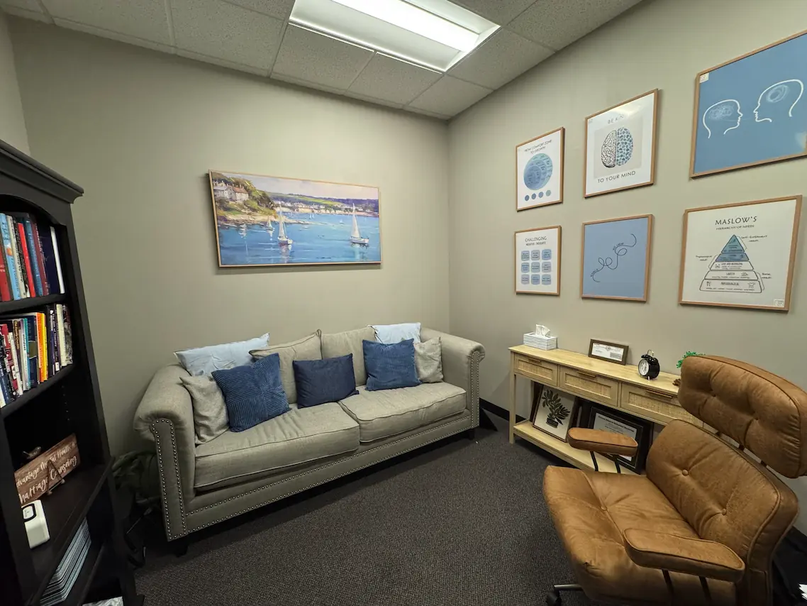 A therapy room at Advantage Mental Health with a sofa, a leather chair, a bookcase, and framed art